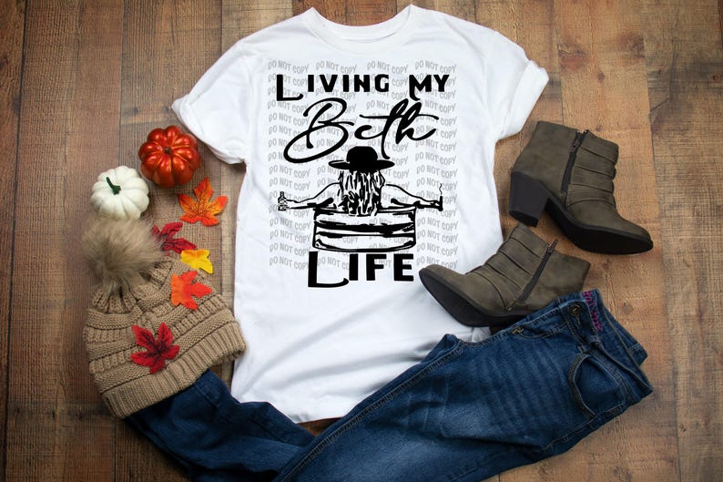 May include: A white short-sleeved t-shirt featuring a black graphic of a person in a hat sitting in a tub with the text "LIVING MY Beth LIFE". The shirt is displayed with a tan knit beanie, blue jeans, and dark brown ankle boots.