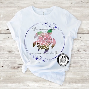 Op de afbeelding: Wit T-shirt met een aquarelontwerp van een schildpad in een cirkel met de tekst "Be comfortable in your own shell". Het ontwerp bevat ook sterren en de tekst "Embroidery Lori Lilley Designs Screenprinting".