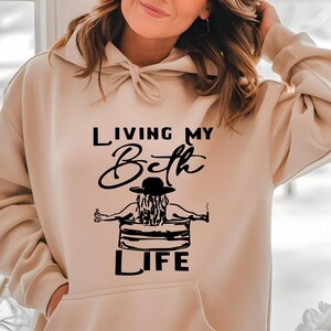 May include: A beige hooded sweatshirt featuring a black graphic print on the front. The design depicts a person in a hat sitting in a tub with the text "LIVING MY Beth LIFE" written in a combination of script and block lettering.