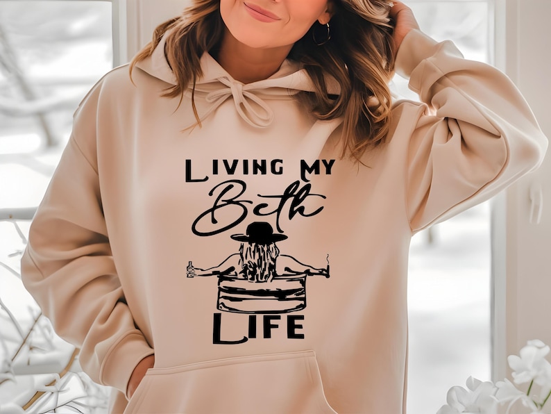 May include: A beige hooded sweatshirt featuring a black graphic print on the front. The design depicts a person in a hat sitting in a tub with the text "LIVING MY Beth LIFE" written in a combination of script and block lettering.