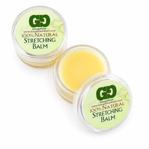 May include: Two small, round, clear plastic containers filled with a pale yellow balm. The lids feature a light green label with the text GaugeGear, GaugeGearBalm.com, 100% Natural Stretching Balm printed in dark green and brown lettering.