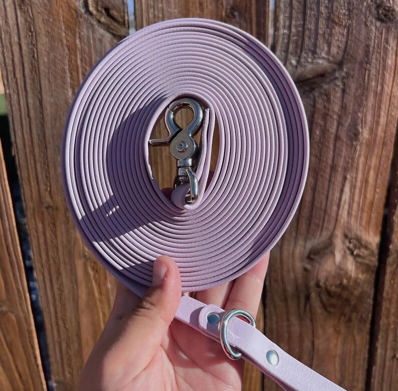 Op de afbeelding: Een opgerolde lavendelkleurige hondenriem met een zilverkleurige metalen sluiting. De riem wordt door een hand vastgehouden, met een bijpassende lavendelkleurige handgreep met een zilveren ring en klinknageldetails. De achtergrond is een houten hek.