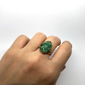 May include: A gold ring with a carved green stone set in the center. The stone is oval shaped and has a leaf design.