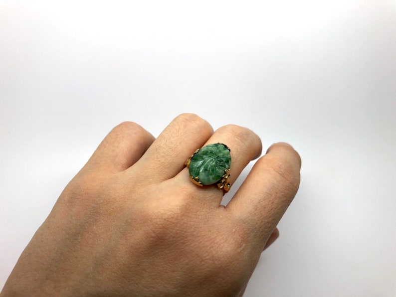 May include: A gold ring with a carved green stone set in the center. The stone is oval shaped and has a leaf design.