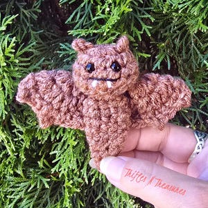 May include: A small, brown, crocheted bat plush toy featuring two black plastic eyes and two white yarn fangs. The bat has textured, spread-out wings and pointed ears, crafted from brown yarn with a visible stitch pattern throughout the body.