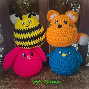 May include: Four handmade crochet animal toys: a bee, a fox, a bunny, and a bird. The bee is yellow and black, the fox is orange, the bunny is pink, and the bird is blue. Each has embroidered facial features. The text "Trifles N Treasures" is at the bottom.