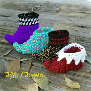May include: Four colorful, crocheted elf shoes in purple, turquoise, red, and black with white accents. The shoes are arranged on a weathered wooden surface with scattered leaves. The text "Trifles N Treasures" is visible.