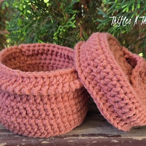 May include: A handmade, dusty rose crochet basket with a matching lid. The basket is round with a slightly flared rim. The lid is also round and fits snugly on top. The text "Trifles N Treasures" is visible in the background.
