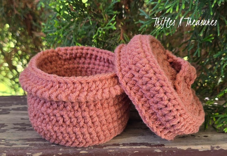 May include: A handmade, dusty rose crochet basket with a matching lid. The basket is round with a slightly flared rim. The lid is also round and fits snugly on top. The text "Trifles N Treasures" is visible in the background.
