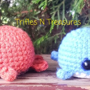 May include: Two handmade crochet plush toys: a coral-colored walrus and a light blue narwhal. The walrus has white tusks and the narwhal has a white horn. Both have black safety eyes. The text "Trifles N Treasures" is in the top left corner.