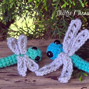 May include: Two crocheted dragonfly toys with teal bodies and light gray wings. The dragonflies feature black bead eyes and are positioned side by side. The text "Trifles N Treasures" is visible in the upper right corner of the image.