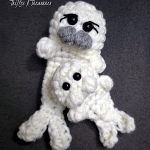 May include: A handmade white crochet seal with a smaller seal attached. The seals have black eyes and gray noses. The text "Trifles N Treasures" is in the upper left corner. The background is a solid gray.