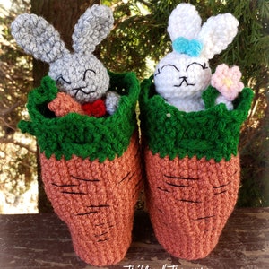 Puede incluir: Dos pantuflas de conejito de Pascua hechas a mano con ganchillo. Un conejito gris con pajarita roja y zanahoria, el otro blanco con lazo azul y flor rosa. Las pantuflas tienen forma de zanahoria con la parte superior verde y el texto "Trifles N Treasures".