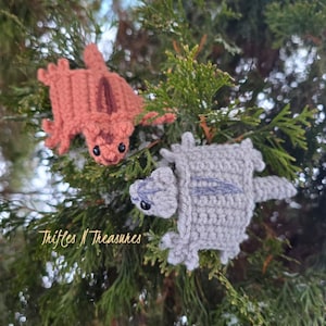 Puede incluir: Dos azucareros de ganchillo hechos a mano en un entorno natural. Uno es melocotón con una raya marrón, el otro es gris con una raya lavanda. Ambos tienen ojos negros y están unidos a ramas de hoja perenne. El texto "Trifles N Treasures" es visible.