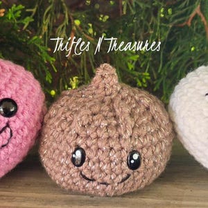 May include: Three crocheted food-themed toys. A pink crocheted strawberry with a smiling face, a brown crocheted onion with a smiling face, and a white crocheted garlic clove with a smiling face. The text "Trifles N Treasures" is in the background.