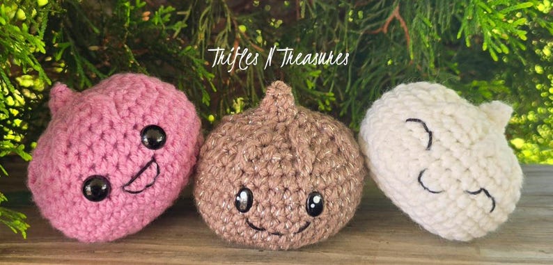 May include: Three crocheted food-themed toys. A pink crocheted strawberry with a smiling face, a brown crocheted onion with a smiling face, and a white crocheted garlic clove with a smiling face. The text "Trifles N Treasures" is in the background.