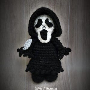 May include: A handmade black and white crochet doll in the shape of the Ghostface mask from the Scream horror movie franchise. The doll has a black hooded robe and a white mask with black eye and mouth details. The text "Trifles N Treasures" is at the bottom.