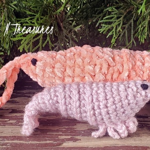 Può includere: Due giocattoli di peluche all'uncinetto a forma di maiali marini rosa e viola. I giocattoli sono realizzati con un filato morbido e peloso e hanno occhi neri. Il maiale marino più grande è rosa e quello più piccolo è viola. I giocattoli sono seduti su una superficie di legno.