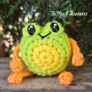 May include: A small, round, crocheted frog plushie featuring a bright lime green body with a yellow circular belly, orange limbs, and large black plastic safety eyes. A stitched black V-shaped mouth is visible. The text Trifles N Treasures is present.