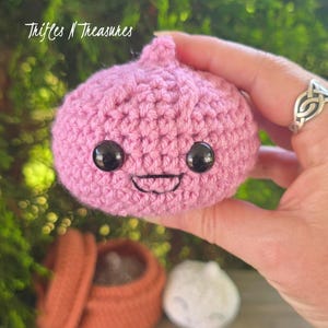 May include: A handmade, pink crocheted onion with black safety eyes and a stitched smile. The onion is held in a hand, with the text "Trifles N Treasures" in the upper left corner. A small, brown container and a white crocheted ball are in the background.