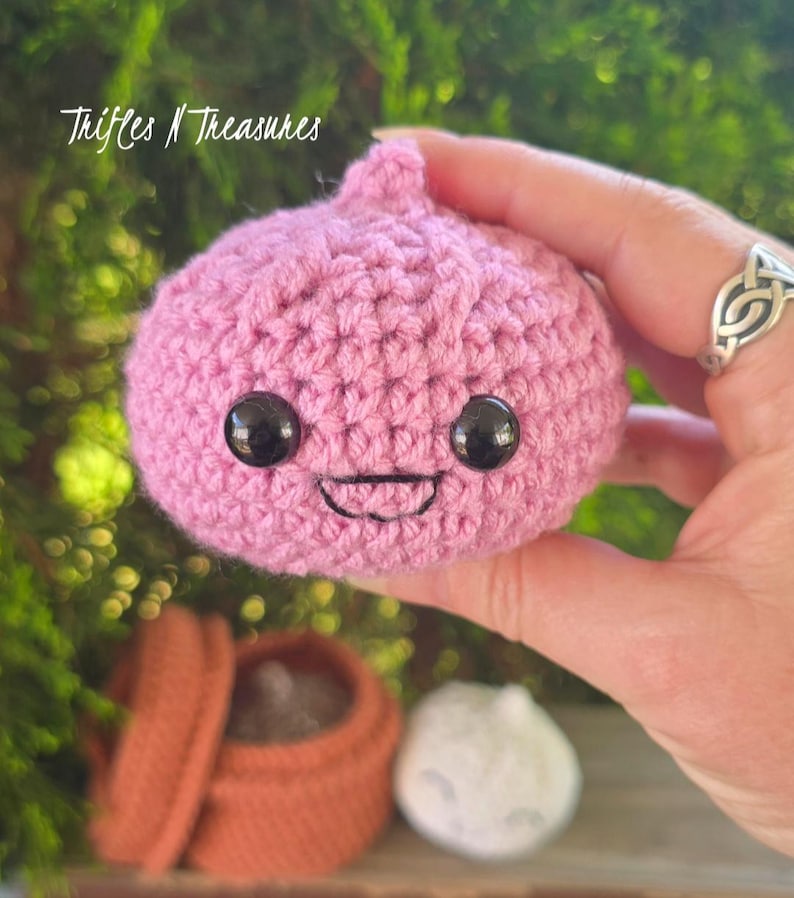 May include: A handmade, pink crocheted onion with black safety eyes and a stitched smile. The onion is held in a hand, with the text "Trifles N Treasures" in the upper left corner. A small, brown container and a white crocheted ball are in the background.