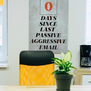 Könnte beinhalten: Ein Poster mit dem Text "0 TAGE SEIT LETZTER PASSIV AGGRESSIVER E-MAIL" hängt an einer Wand. Darunter steht ein schwarz-gelber Bürostuhl vor einem Holztisch. Auf dem Schreibtisch steht eine Topfpflanze.