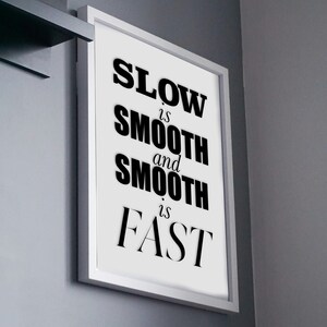 May include: A framed print with the text "SLOW is SMOOTH and SMOOTH is FAST" in bold black letters. The white framed art is mounted on a gray wall. A Google Pixel phone box sits on a shelf above the frame.