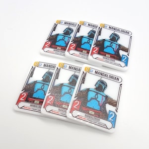 May include: Five rectangular white game tokens featuring a graphic illustration of a Mandalorian character in blue armor. Each token displays the text MANDALORIAN, a red number 2, a blue number 2, and the text SHIELDED TOKEN UNIT GROUND.