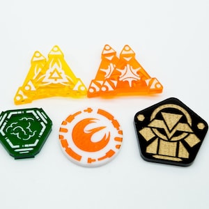 Op de afbeelding: Vijf acryl-tokens met verschillende ontwerpen. De tokens zijn in verschillende vormen, waaronder een driehoek, een cirkel en een vijfhoek. De tokens zijn in verschillende kleuren, waaronder groen, oranje, geel en zwart.