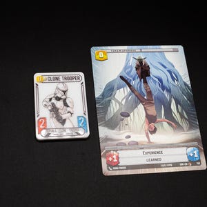 K&ouml;nnte beinhalten: Zwei Star Wars-Spielkarten auf schwarzem Untergrund. Eine Karte zeigt eine Clone Trooper-Illustration, die andere eine Szene mit Yoda und einer Person, die einen Handstand macht. Die Karten haben Text und Spielstatistiken.