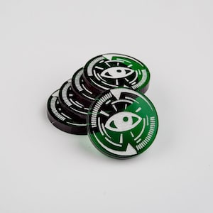 Op de afbeelding: Een stapel van zes ronde, donkergroene en zwarte tokens met een wit oogsymbool in het midden. De tokens hebben een cirkelvormig ontwerp rond het oog. De achtergrond is effen wit.