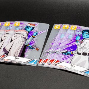 Könnte beinhalten: Zwei Sammelkarten mit einem blauhäutigen Charakter in einem weißen Mantel, der eine Waffe hält. Die Karten haben einen lila und silbernen Hintergrund mit dem Wort "Spy" und der Zahl "2". Die Karten sind für ein Spiel.
