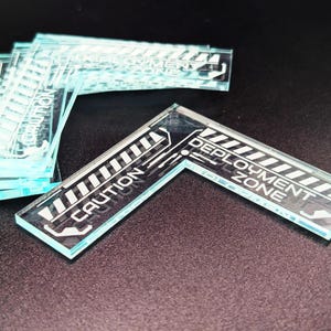 May include: A stack of L-shaped clear acrylic markers featuring white printed graphics. Each marker displays the text CAUTION and DEPLOYMENT ZONE alongside diagonal striped patterns. The design is etched or printed on the transparent material.