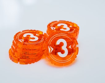 Pode incluir: Uma pilha de fichas de jogo de plástico laranja com o número "3" impresso em cada uma. As fichas estão empilhadas sobre uma superfície branca.