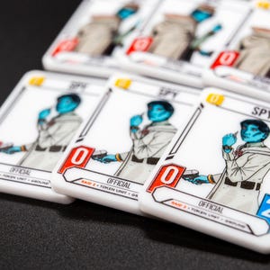 K&ouml;nnte beinhalten: Wei&szlig;e Spielkarten mit einem blauh&auml;utigen Charakter in heller Uniform, beschriftet mit "SPY" und "OFFICIAL". Die Karten haben rote und blaue Akzente, mit einer Null oder Zwei in der Ecke. Die Karten liegen auf einer schwarzen Oberfl&auml;che.