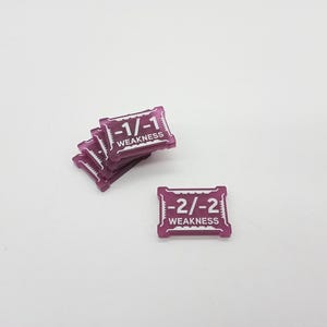 May include: A collection of small, rectangular purple game tokens with white text. One token displays -2/-2 WEAKNESS, while a stacked group of tokens displays -1/-1 WEAKNESS. The tokens feature a notched border design and a translucent finish.