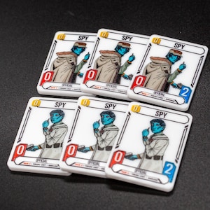 May include: Six white game cards featuring a blue-skinned character illustration, labelled "SPY". The cards have a red or blue number, and the number "0" is on most cards. The cards are on a black surface.