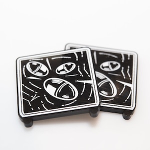 May include: Two black and white square coasters with a graphic design of three stylized eyes. The coasters have a black background and white lines and circles.