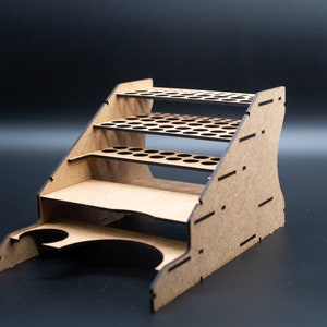 Puede incluir: Un soporte de madera para botes de pintura con cinco niveles de agujeros circulares para guardar botes de pintura. El soporte tiene una base curva con una pequeña taza para guardar pinceles.