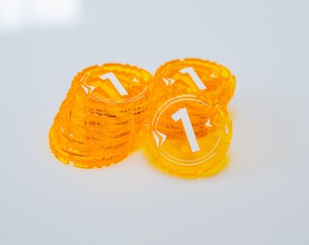 Pode incluir: Várias pilhas de fichas de jogo laranja transparentes, cada uma exibindo o número 1 em branco. As fichas circulares têm uma borda entalhada e um design de seta branca. Estão dispostas sobre uma superfície branca.