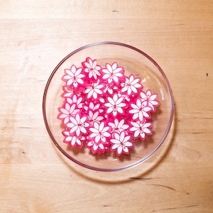 Puede incluir: Un bol de vidrio transparente lleno de pinzas para el pelo en forma de flores rosas con centros blancos.