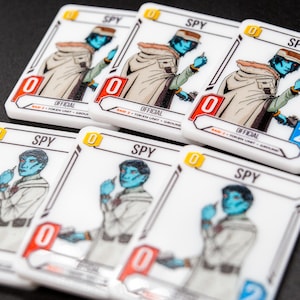 K&ouml;nnte beinhalten: Eine Sammlung wei&szlig;er Spielkarten mit einem blauh&auml;utigen Charakter in einem beigefarbenen Mantel, der mit "SPY" beschriftet ist. Jede Karte hat einen Wert von "0" oder "2" und das Wort "OFFICIAL". Die Karten sind auf einer dunklen Oberfl&auml;che angeordnet.