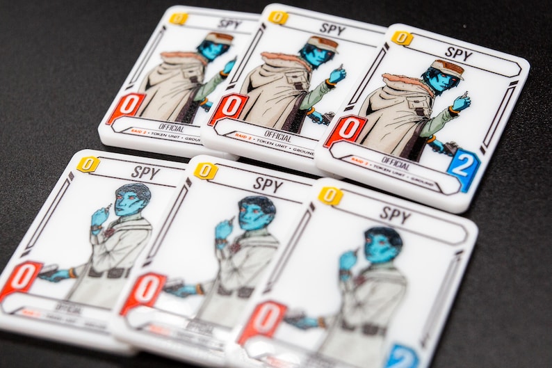K&ouml;nnte beinhalten: Eine Sammlung wei&szlig;er Spielkarten mit einem blauh&auml;utigen Charakter in einem beigefarbenen Mantel, der mit "SPY" beschriftet ist. Jede Karte hat einen Wert von "0" oder "2" und das Wort "OFFICIAL". Die Karten sind auf einer dunklen Oberfl&auml;che angeordnet.