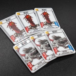 May include: Five rectangular game cards with white borders and black text. Three cards feature a red, tentacled creature with a central eye, labelled BEAST. Two cards feature a grey, multi-legged creature with sharp teeth, also labelled BEAST.