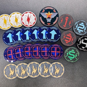 Puede incluir: Una colección de fichas de juego circulares con varios diseños, incluyendo rayos, flechas hacia arriba, iconos numerados del uno al cinco, un emblema de estrella central y símbolos abstractos de llamas en colores rojo, azul, amarillo, verde y negro.