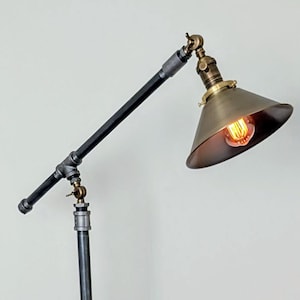 May include: Industrial-style floor lamp with a black metal frame and a brass-colored lampshade. The lamp features a vintage Edison bulb and a unique pipe fitting design. The lamp stands on a four-legged base and is plugged into a wall outlet.