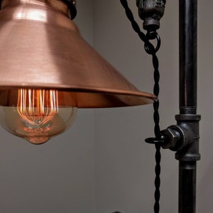 May include: Close-up of an industrial-style lamp with a copper shade and exposed filament bulb. The lamp features a black twisted cord and a metal pipe frame. The warm glow of the bulb contrasts with the cool tones of the metal.