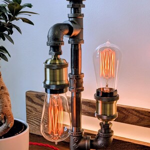 May include: Industrial-style desk lamp made from black metal pipes and fittings, featuring two Edison-style light bulbs with visible filaments. The lamp has a red valve handle on top and sits on a wooden base. The warm light creates a cozy ambiance.