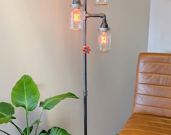 Könnte beinhalten: Industrielle Stehlampe mit schwarzem Rohrrahmen und vier Gläsern als Lampenschirme. Die Lampe hat Edison-Glühbirnen und ein rotes Ventil. Der Sockel besteht aus Rohren. Die Lampe steht in der Nähe einer Topfpflanze und eines braunen Lederstuhls.