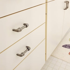 May include: Close-up of white cabinets with industrial-style pipe drawer pulls. The metal pulls are a dark gray color and have a unique, curved design. The cabinets are clean and modern, with a minimalist aesthetic.